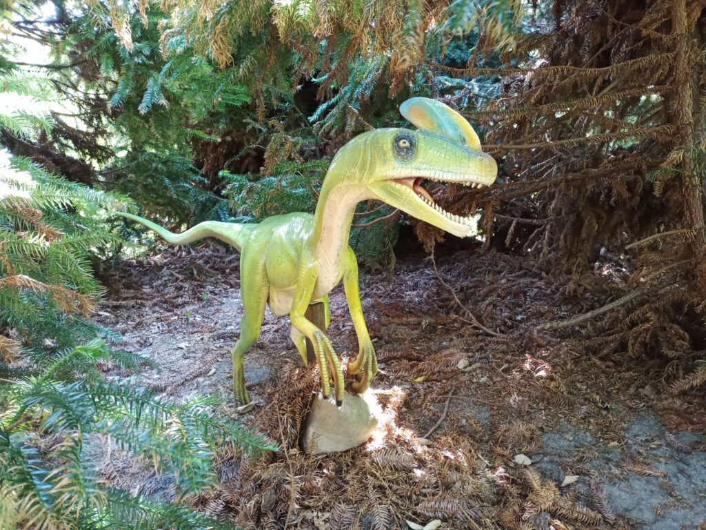Een lichtgroene dinosaurus staat op de grond tegen een achtergrond van dennenbomen. 
