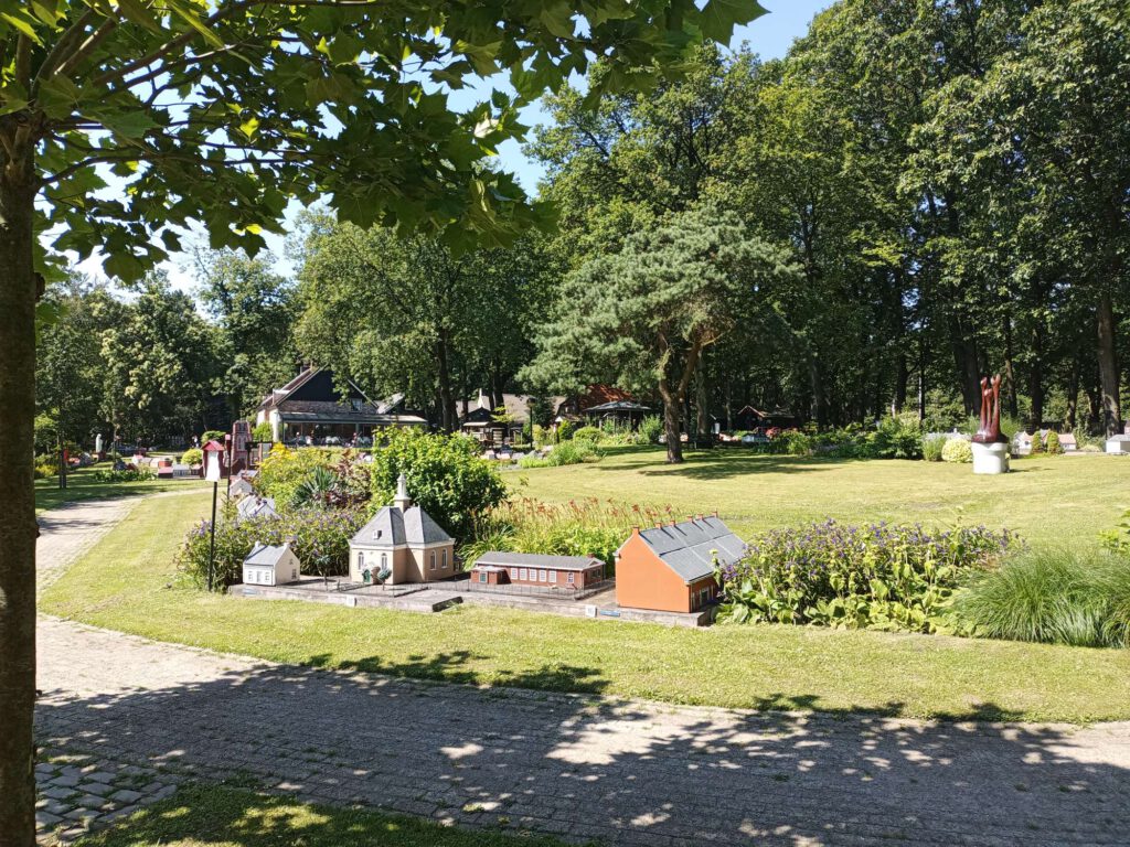 Je kijkt naar een grasveld waar bomen staan, Langs de paden bevinden zich miniatuur huizen met daarachter lage begroeiing van planten en bosjes.