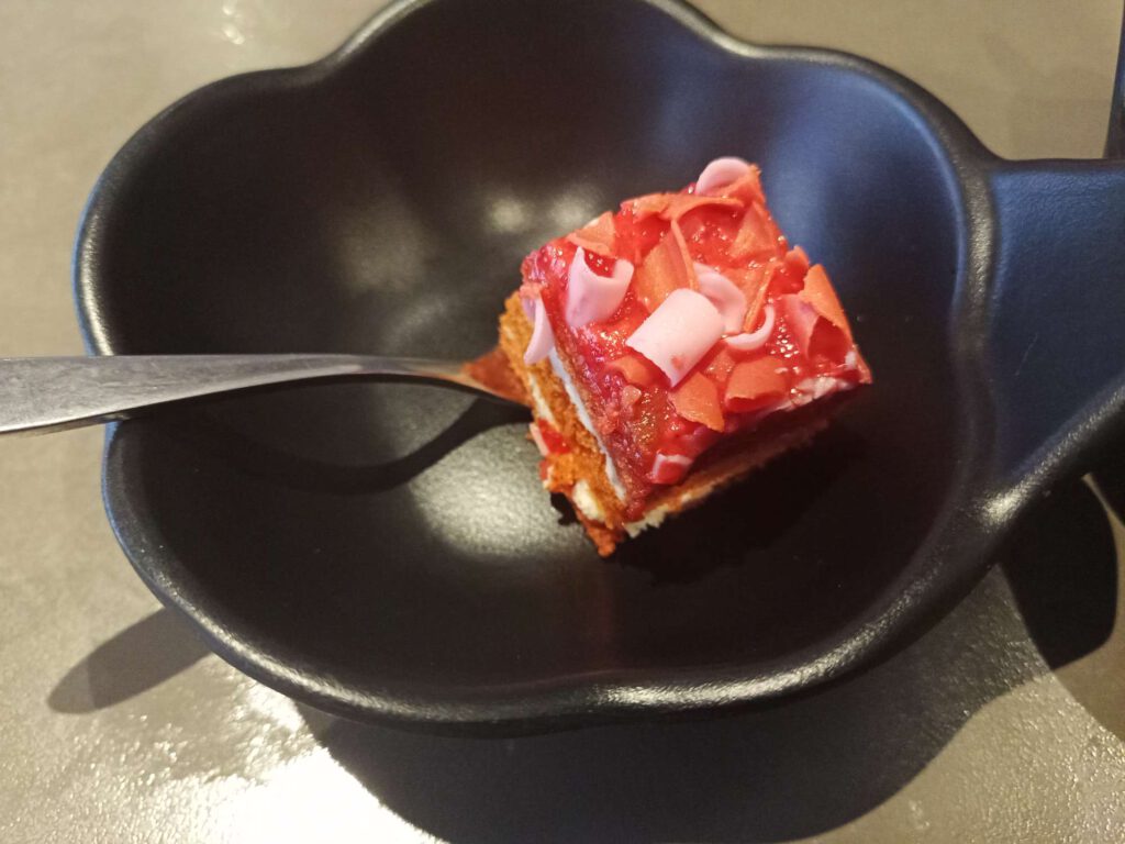 Op een vorkje ligt een stukje red velvet cake wat wat rood-roze vlokken heeft en niet veel groter is een kubus van 2 cm.