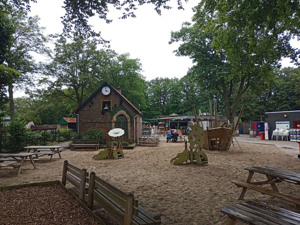 Een speeltuin van attractiepark De Waarbeek met verschillende houten speeltoestellen en een schip. Daarachter een horecapunt.