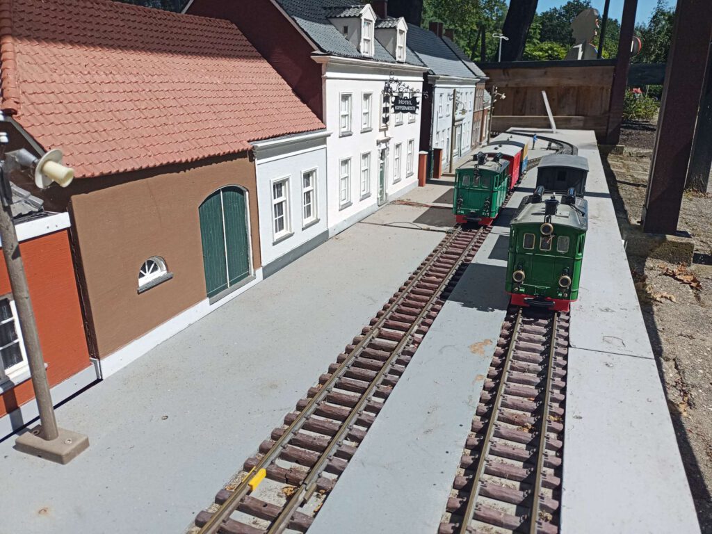 Links staan miniatuur huizen. Je kijktuit naar twee sporen naast elkaar, op elk spoor staat een miniatuurtram in het groen. Het rijdt op een grijze ondergrond.