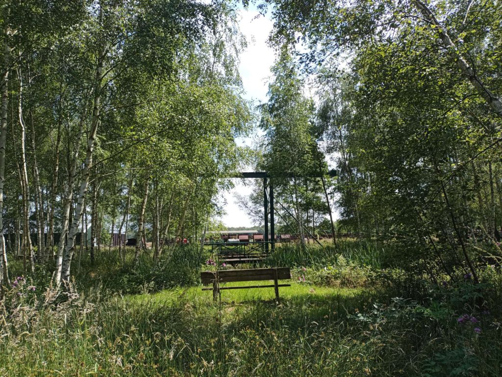 Op de foto zie je een bankje staan centraal in het groen tussen bomen. Je kijkt er tussendoor naar een groene ophaalbrug en op de achtergrond staan kiepwagonnen. 