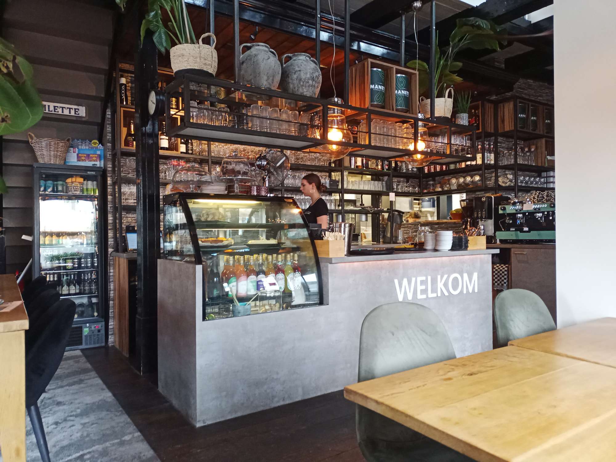 Een grijze bar met Welkom erop, daar boven een vitrine met gebak, een koffie automaat en diverse likeuren en glazen.
