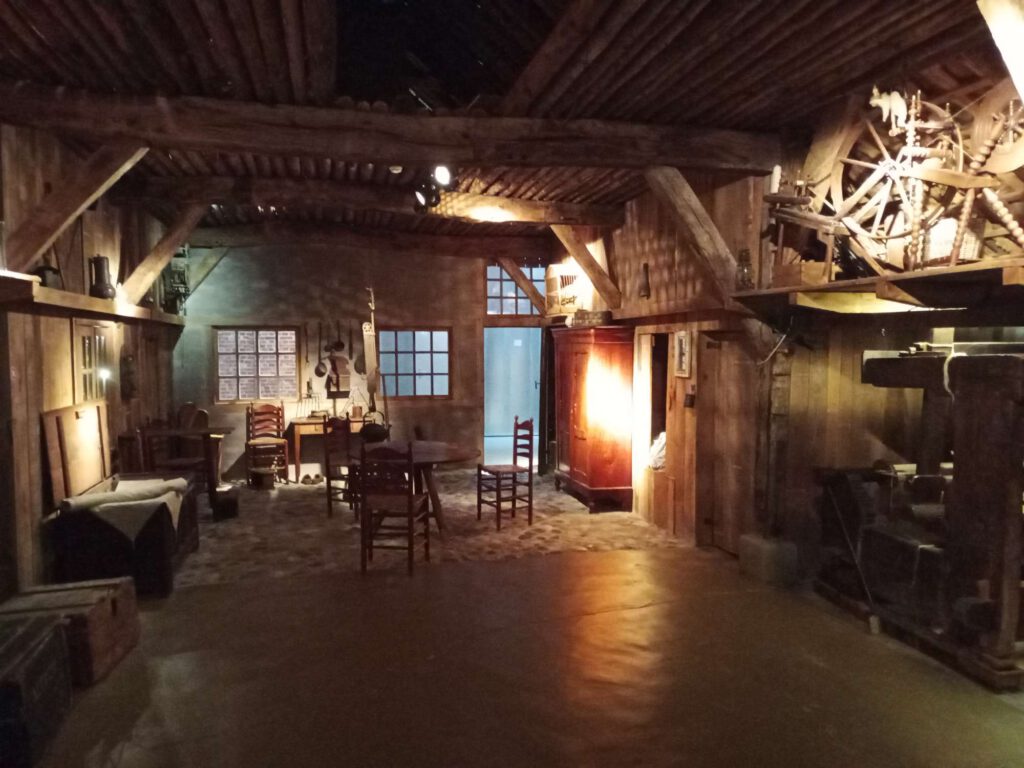 De boerderij heeft een houten afwerking in de wanden. Links staan kratten en kisten, linksachter een gelegenheid tot koken. In het midden een houten tafel met houten stoelen erom. Rechtsachter is een bedstee, rechts zie je een textiel weefgetouw en daarboven een spinnewiel.