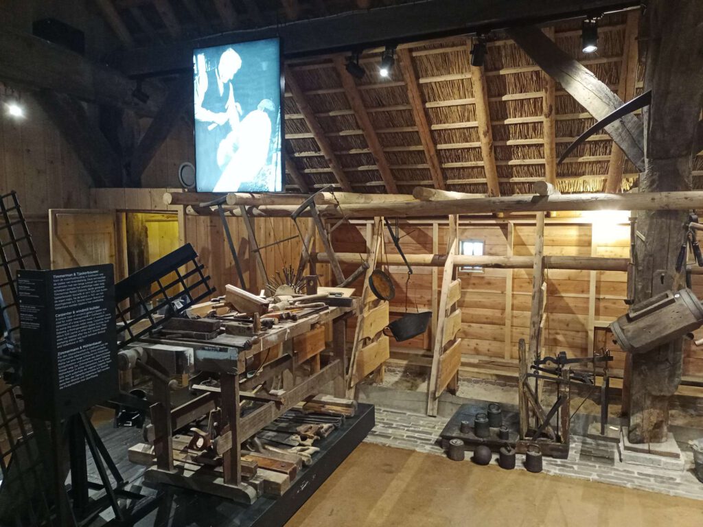 Tegen de wand staan houten schotten om het vee uit elkaar te houden. Daarvoor staat een houten werkbank met verschillend materialen. Rechts een soort weegschaal met gewichten ervoor.