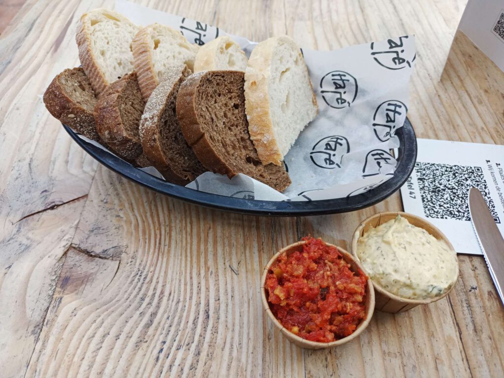 In een zwart mandje met papier erin liggen 8 stukjes brood. Daarvoor staan twee kuipjes, links met tomatentapenade en rechts met kruidenboter. Op de tafel ligt een mes wijzend naar de QR code van de tafel.