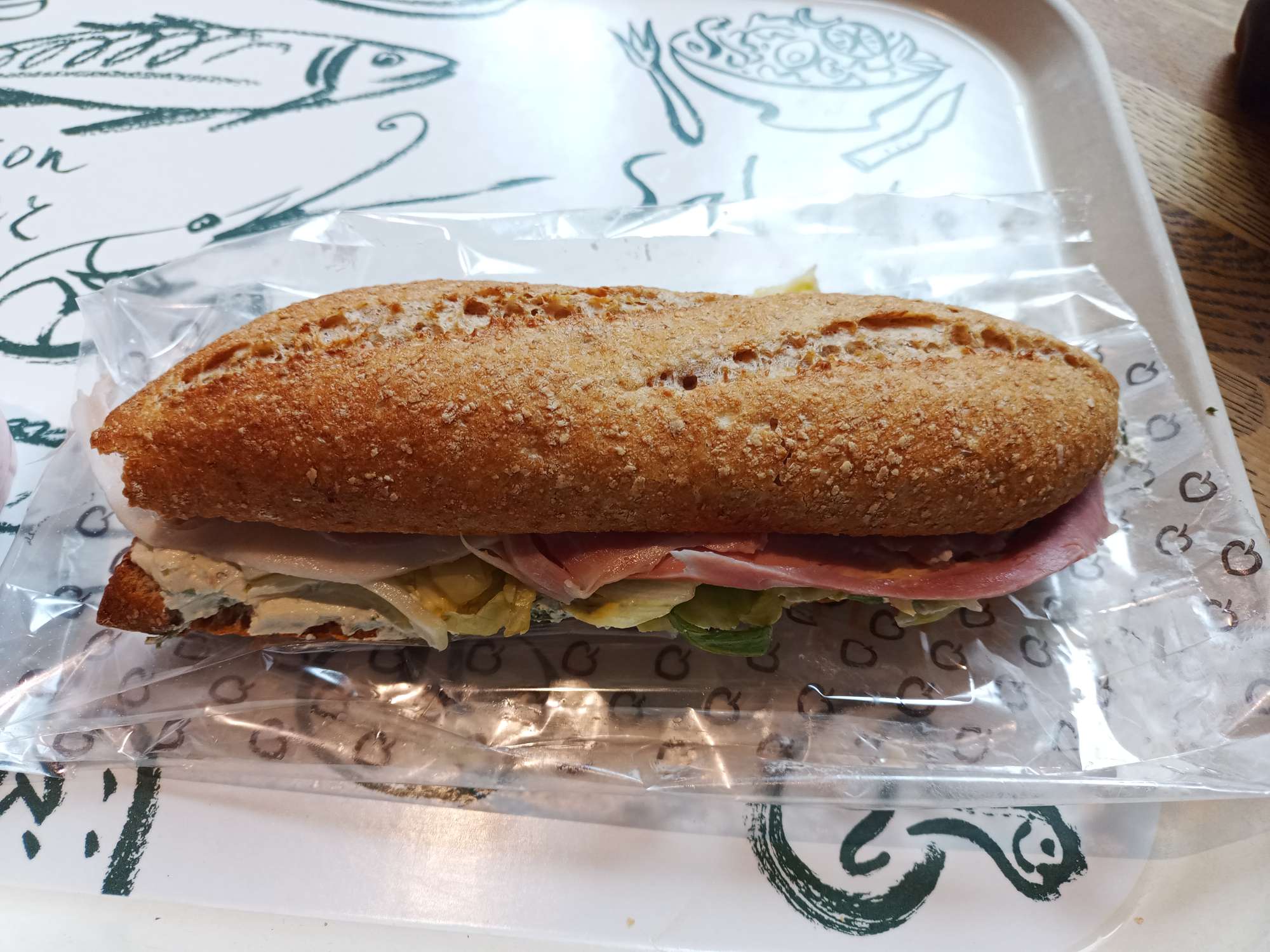 Een bruin broodje met roomkaas en ham op een plastic zakje op een dienblad van la place.