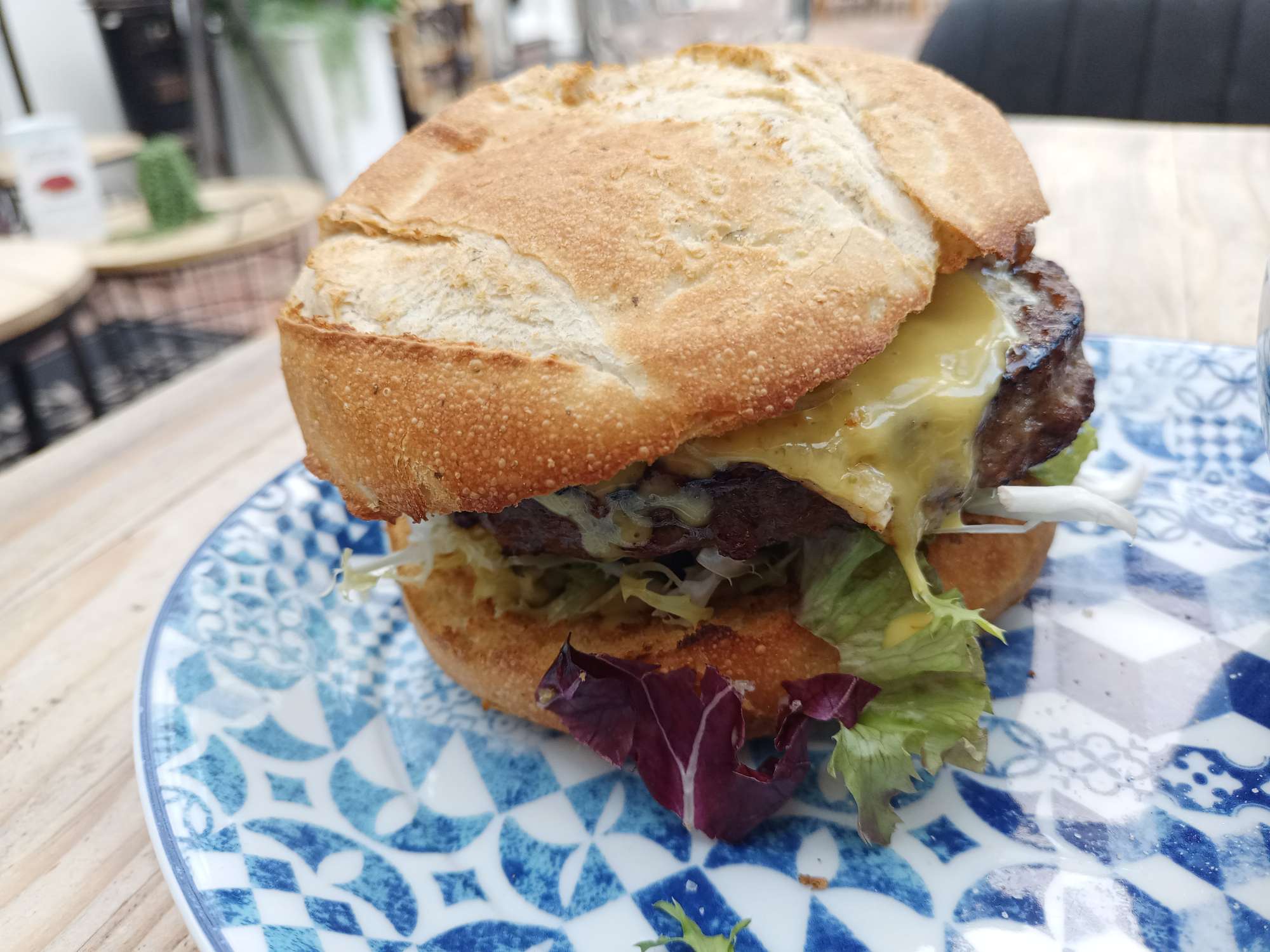 Op een delfsblauw bordje ligt een hamburger. De hamburger ligt op een italiaanse bol, met sla, tomaat en een runder hamburger waar kaas en saus naar beneden druipt.