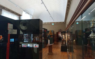 De zaal boven in het Universiteitsmuseum, rechts zijn glazen vitrines, links zijn verschillende objecten te zien in vitrines. Het plafond is wit. In de vitrines links zijn ook nog bewegende beelden.