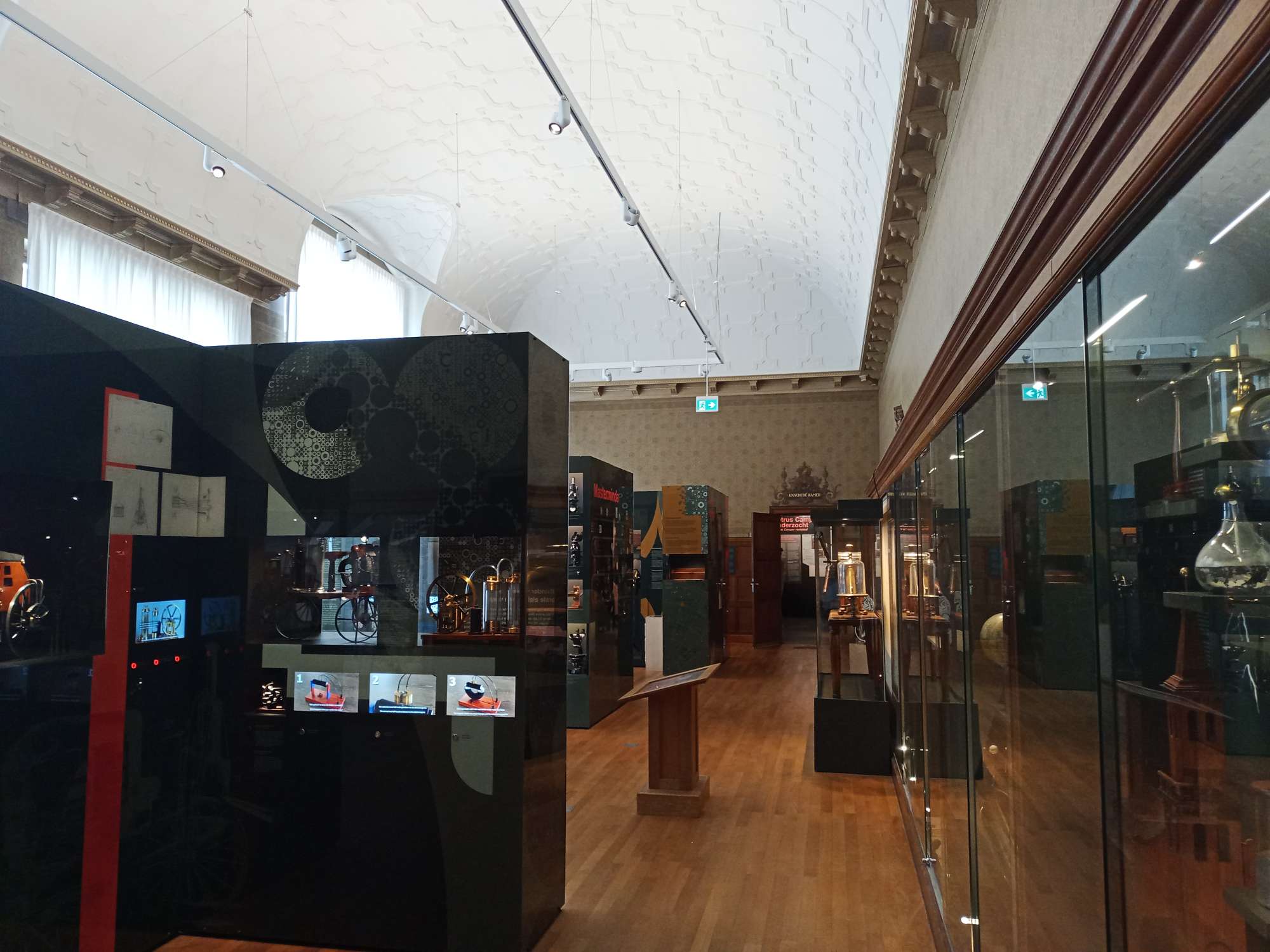 De zaal boven in het Universiteitsmuseum, rechts zijn glazen vitrines, links zijn verschillende objecten te zien in vitrines. Het plafond is wit. In de vitrines links zijn ook nog bewegende beelden.