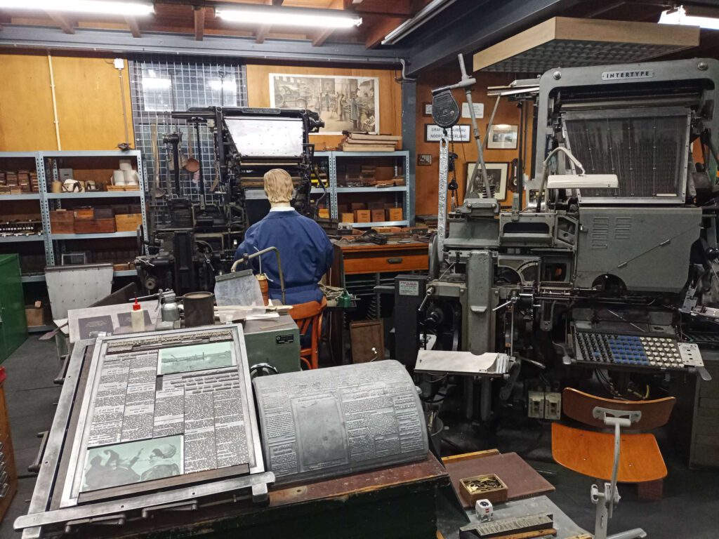 Rechts staat een intertype machine. Deze heeft aan de bovenkant het alfabet in loden letters staan. Daaronder zit een toetsenbord met de letters. Een typist toets de letters van een woord in, waarna er een zin ontstond die in een soort houten bak (een zetraam) werd gezet om te kunnen drukken. Later werden de grote stukken in 1 keer in lood gezet, waar links op de foto voorbeelden van zijn, in een setraam en in een halve rol voor een pers.