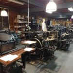 Er staan diverse drukmachines in de museumdrukkerij, voornamelijk van bruin en zwarte kleuren. Daarboven hangen witte lampen.