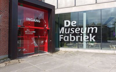 Links is een glazen deur met een rode achterwand. Rechts staat op het glas De Museum Fabriek.