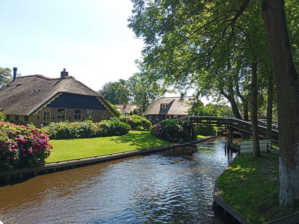 Links van de foto is een boerderij te zien, verscholen achter bosjes en gras. Daarvoor lang van links onder naar rechtsboven loopt een kanaal. Rechts loopt een brug over het kanaal. Rechts is ook de waterkant nog te zien met gras en bomen. 