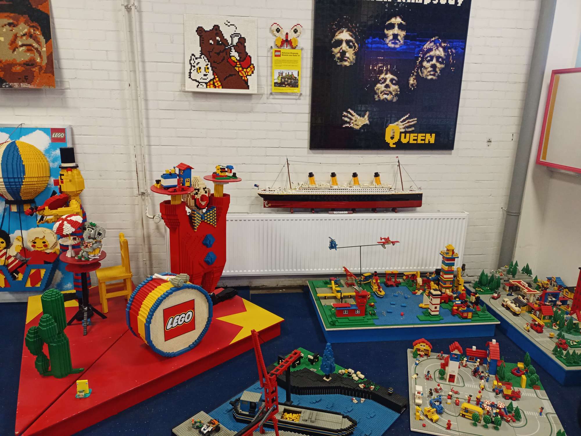 Op de foto staan verschillende bouwwerken van Lego en het lego logo. De bouwwerken die hier te zien zijn onder andere een portret van Queen, een trommel, een clown, een boot in een vaargeul en de titanic.