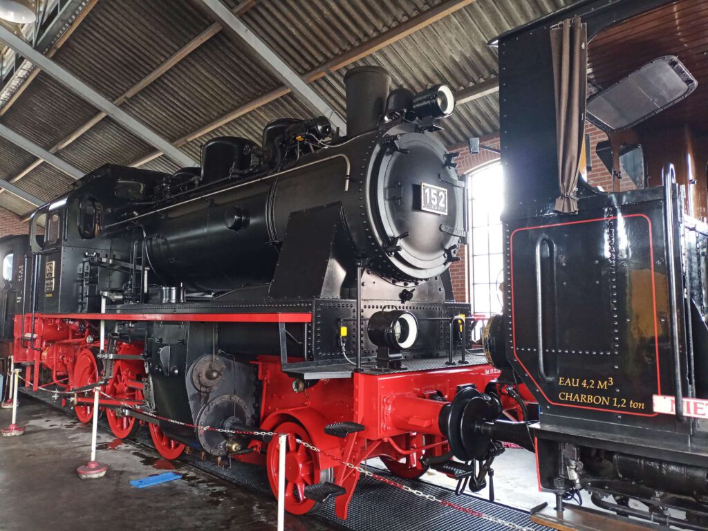 Deze locomotief in de remise heeft een rood onderstel en de ketel is zwart. De locomotief heeft drie assen.