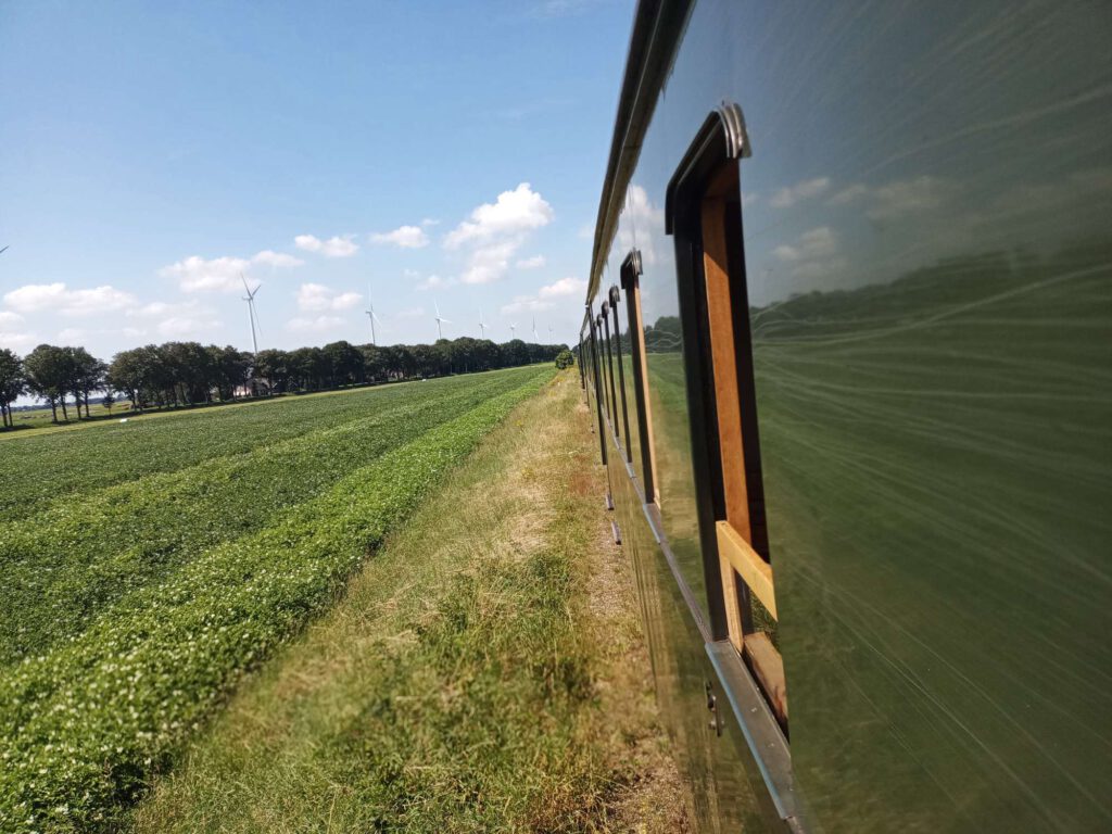 Rechts van de afbeelding zie je de trein en rijtuigen in het groen. Links, naast het spoor zie je de groene struiken van aardappelgewas en in de verte windmolens. De hemel is blauw met enkele witte wolken.