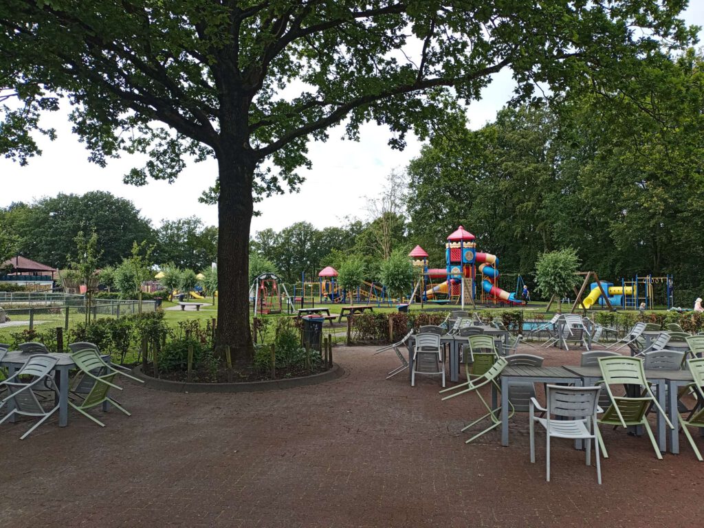 Op de voorgrond een terras waar de stoelen tegen de tafels staan vanwege het regenachtige weer. Daarachter staan in rode, blauwe, oranje, gele speel- en klimtoestellen. Er is tevens een rolstoelschommel.