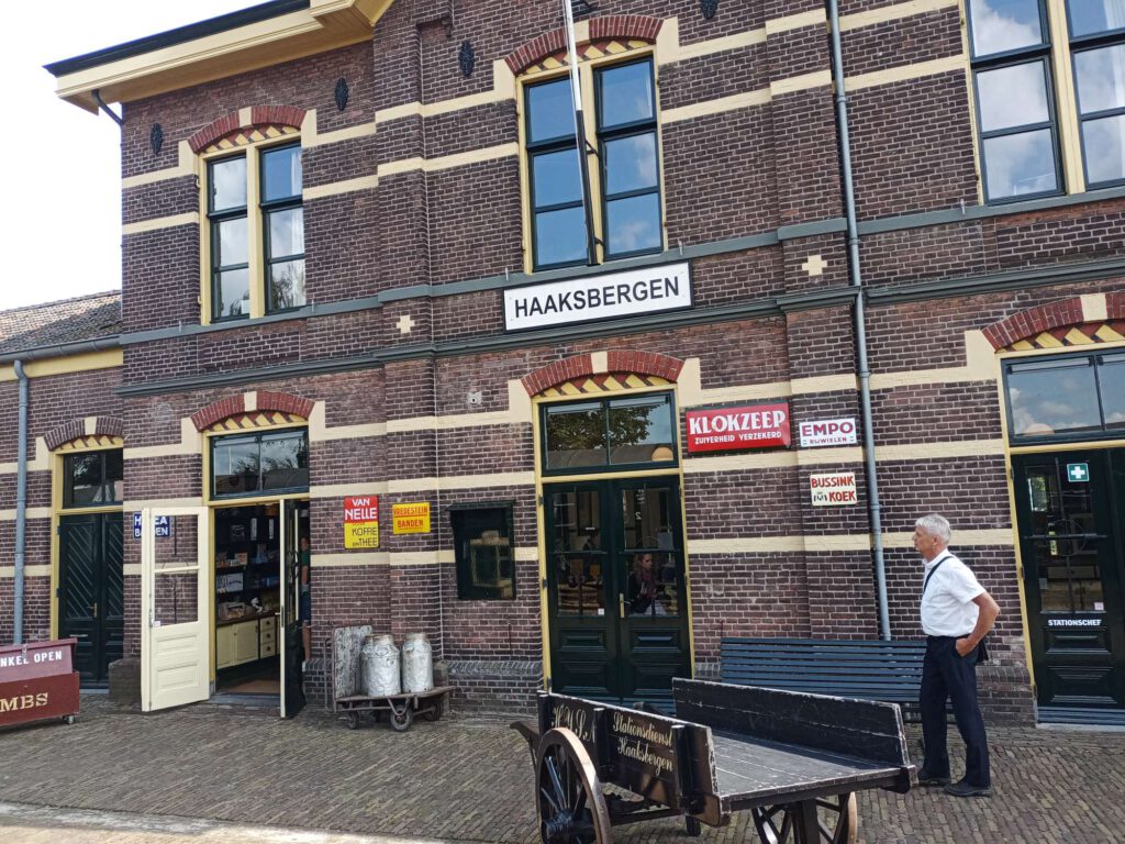 Je ziet hier een stationsgebouw van rode en gele stenen. Hier staat Haaksbergen op, met verschillende reclameborden. Voor het stationsgebouw staat een conducteur en een handkar om dingen mee te vervoeren.