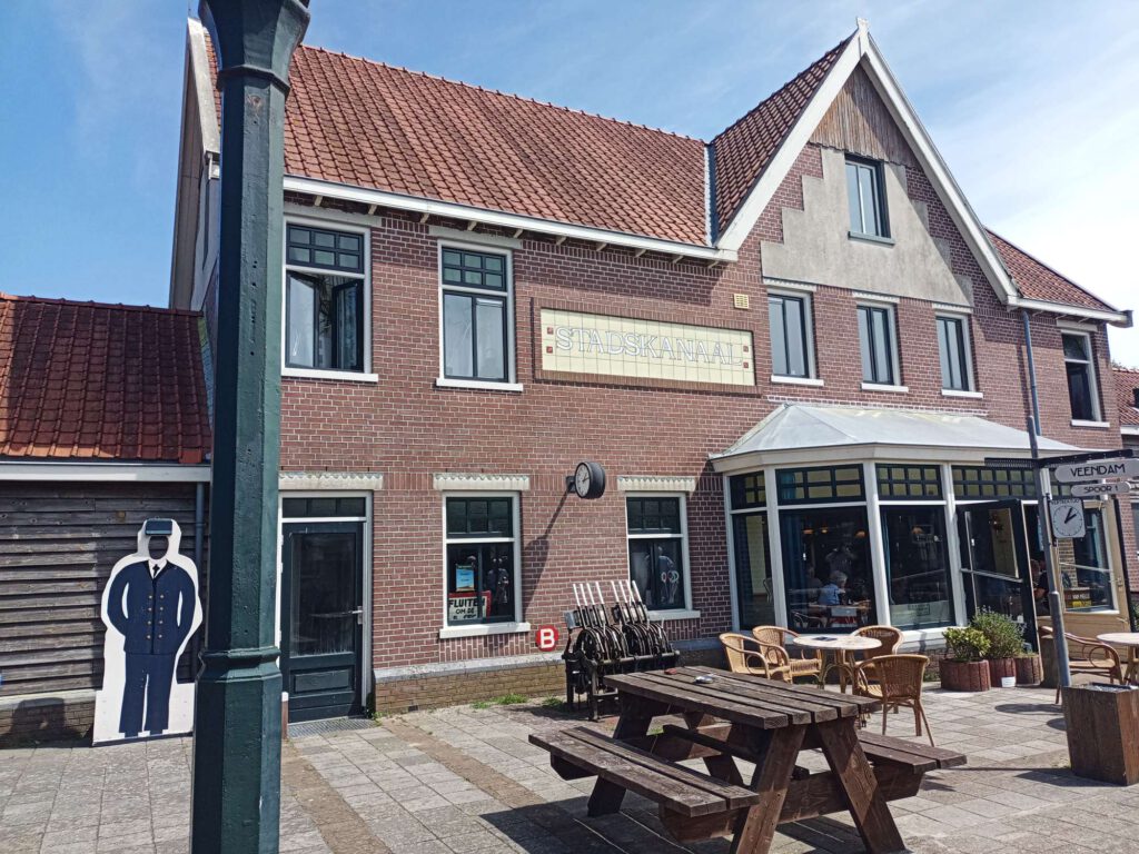 De buitenkant van station Stadskanaal zit in een soort gebouw et een puntdak, waar met lichte letters op staat Stadskanaal. Voor het gebouw staat een terras. Rechts is een klok te zien met een vertrektijd.