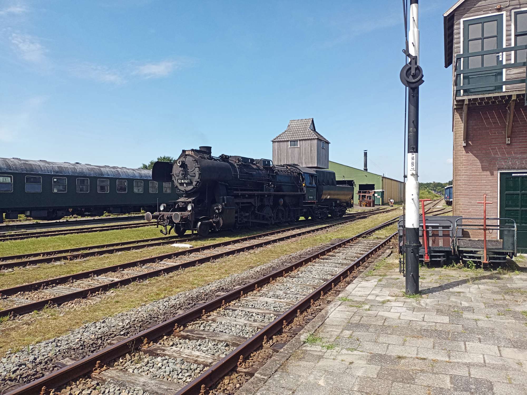 Een zwarte stoomlocomotief met vijf assen. Links staan nog rijtuigen op de rails. rechts is een seinhuis te zien.