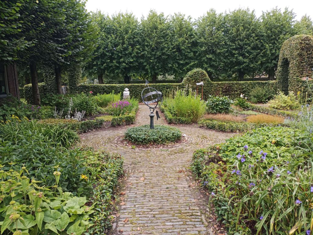 De tuin heeft laag groen, links zijn wat gele bloemen, rechts blauwe. In het midden van het pad staat een metalen beeld van een bol met een pijl er door. Achteraan zijn nog wat paarse bloemen en een heg met bogen. Tegen de achtergrond staan bomen.