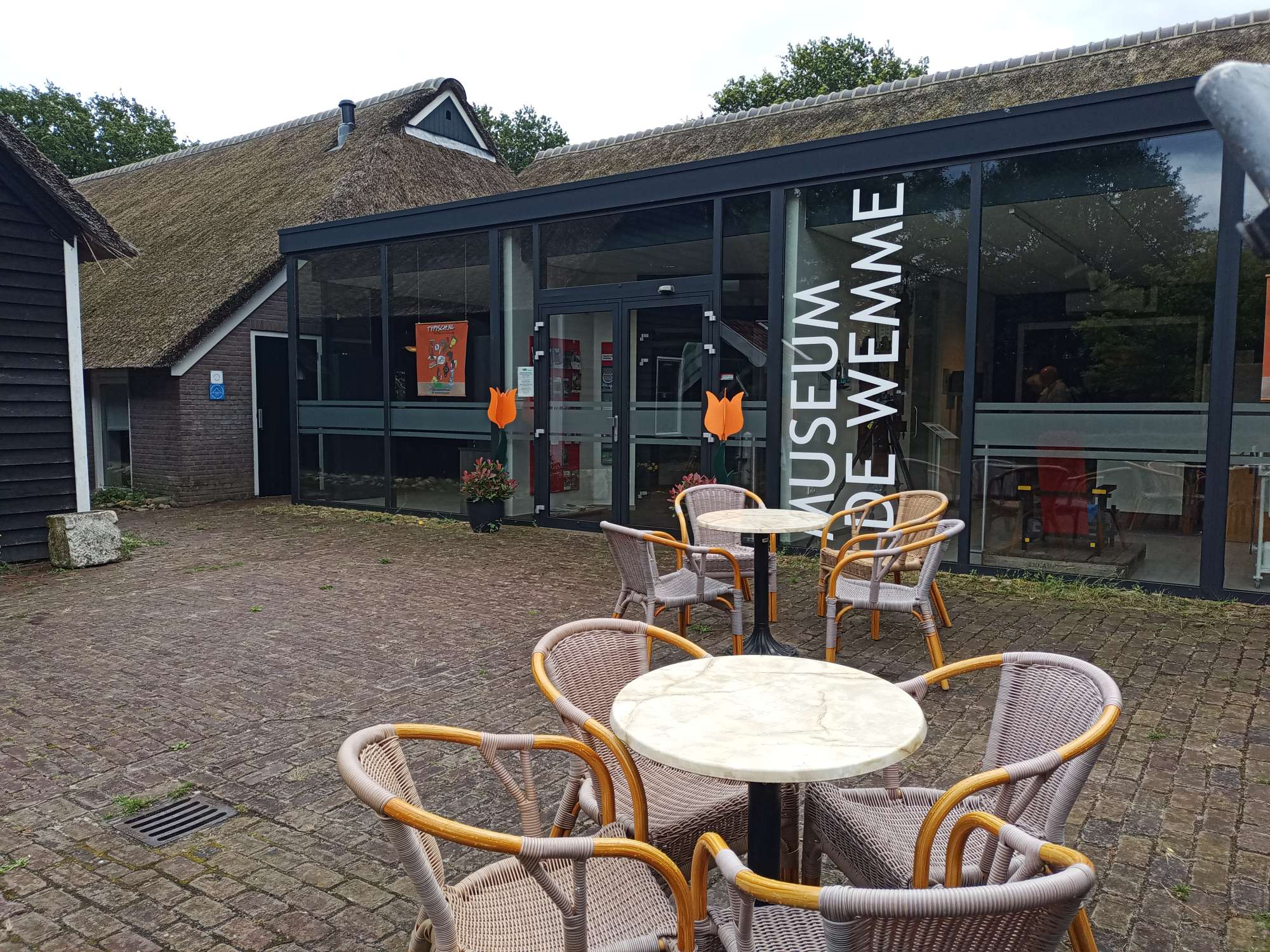 Op de voorgrond staat het terras met tafels en rieten stoelen. Op de achtergrond is de voorgevel van glas te zien met daarop in witte letters De Wemme.