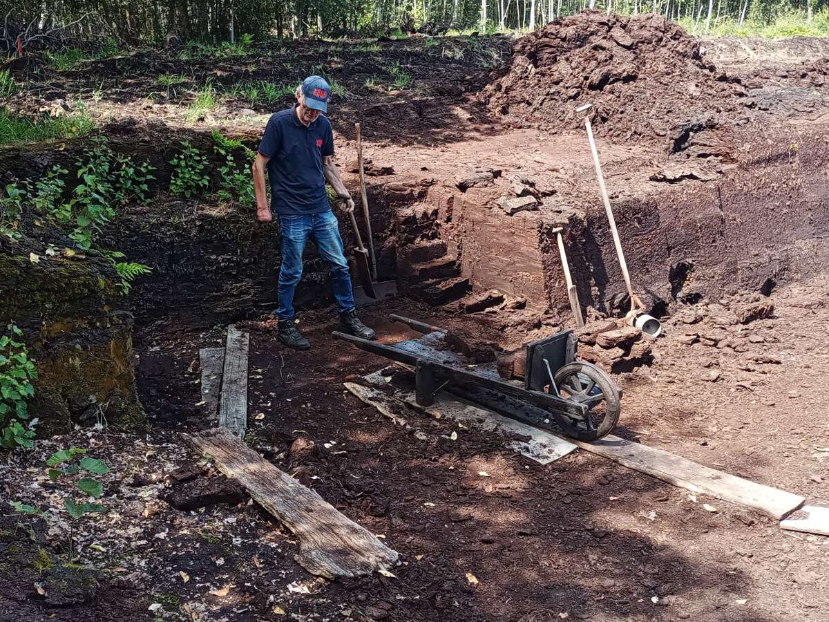 Een medewerker van het veenpark met een blauwe spijkerbroek en een blauw T-shirt en een pet, staat op het veen vooraf het stuk waar nog afgestoken is. Voor hem staat een kruiwagen waar verschillende stukken veen op een kruiwagen staan. Rondom de medewerker staan verschillende gereedschappen, zoals een mes om het turf af te steken.