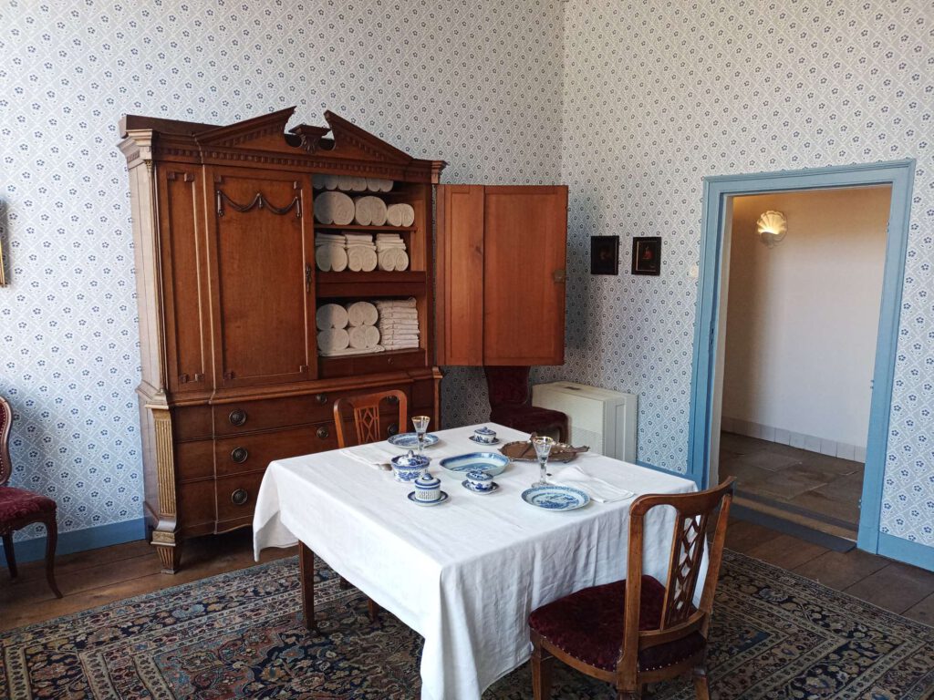 De eetkamer van het Palthe Huis. Tegen de achter wandstaat een kast met linnen. Daarvoor staat een vierkante tafel  met houten stoelen en een rood beklede zitting. Op tafel staat servies in Delfsblauw en zilver bestek.