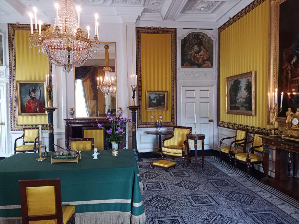 Er hangt een kroonluchter aan het plafond. De wanden zijn wit met gele en gouden accenten. Er staat een tafel met een groen tafelkleed. De stoelen zijn van bruin hout en de zitting is geel bekleed.