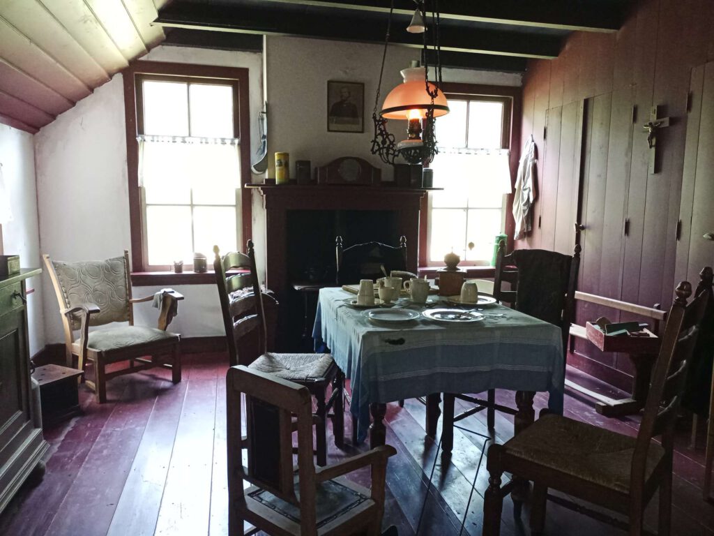 Op de foto is de eetkamer te zen. Er is een houten tafel in het midden met een blauw tafelkleed welke gedekt is. Daar om heen staan stoelen. Tegen de achterwand is de kachel te zien, rechts de beddenstee. Boven de eettafel brand een lamp op elektra.