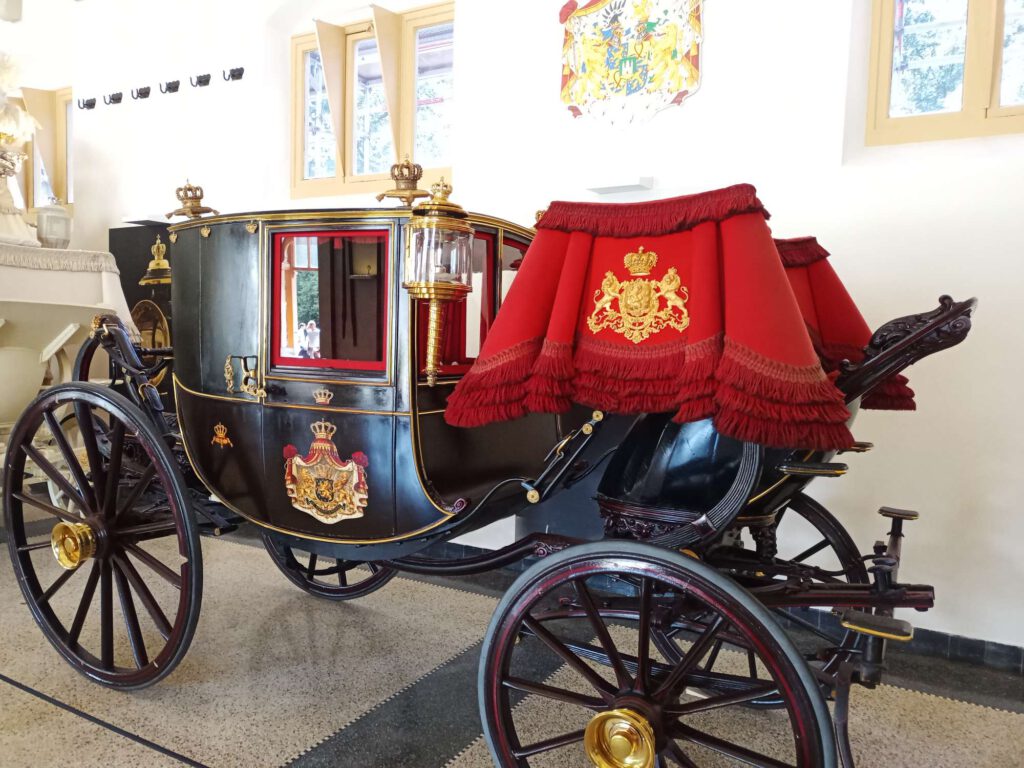 Op de foto staat een zwarte koets. De spaken van de wielen zijn zwart, het ronde deel donkerrood en de wieldoppen goudkleurig. De zitting van de koetsier is bekleed met een rode stof met een gouden embleem van twee leeuwen. Op de koets staat hier ook een afbeelding. De koets heeft gouden accenten aan de onderkant en bovenkant van de koets.