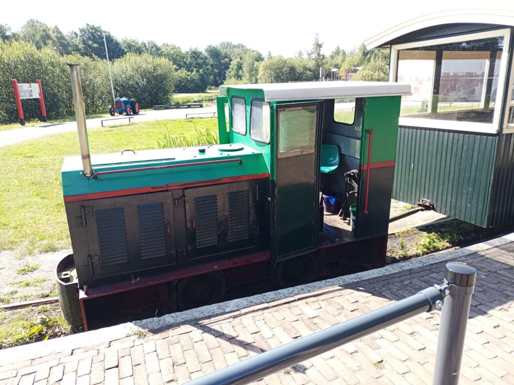 Een locomotief staat voor de wagons. De locomotief is donkergroen van kleur toto halverwege, daar is een rode streep en daarboven lichtgroen. Bij de machinist is het dak nog wit. Achter de locomotief zijn wagons van hout met witte kozijnen en ramen erin, waar passagiers in mee kunnen.