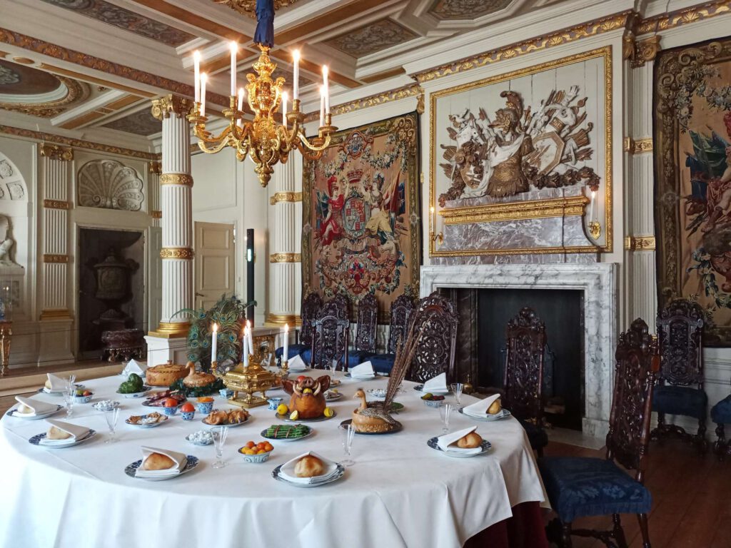 Een gouden kroonluchter, met daaronder een goed gevulde eettafel. Het tafelkleed is wit, daarop staat servies en diverse etenwaren.