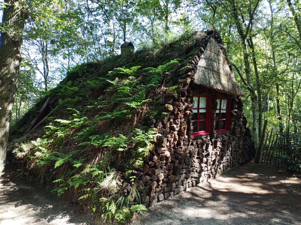 In een bos staat een plaggenhut. Dit is gemaakt van afgestoken stukkengrond waar dan nog wel aan de voor- en achterkant ramen zitten, deze hebben rode kozijnen. Op de plaggenhut zelf  groeien ook weer planten.