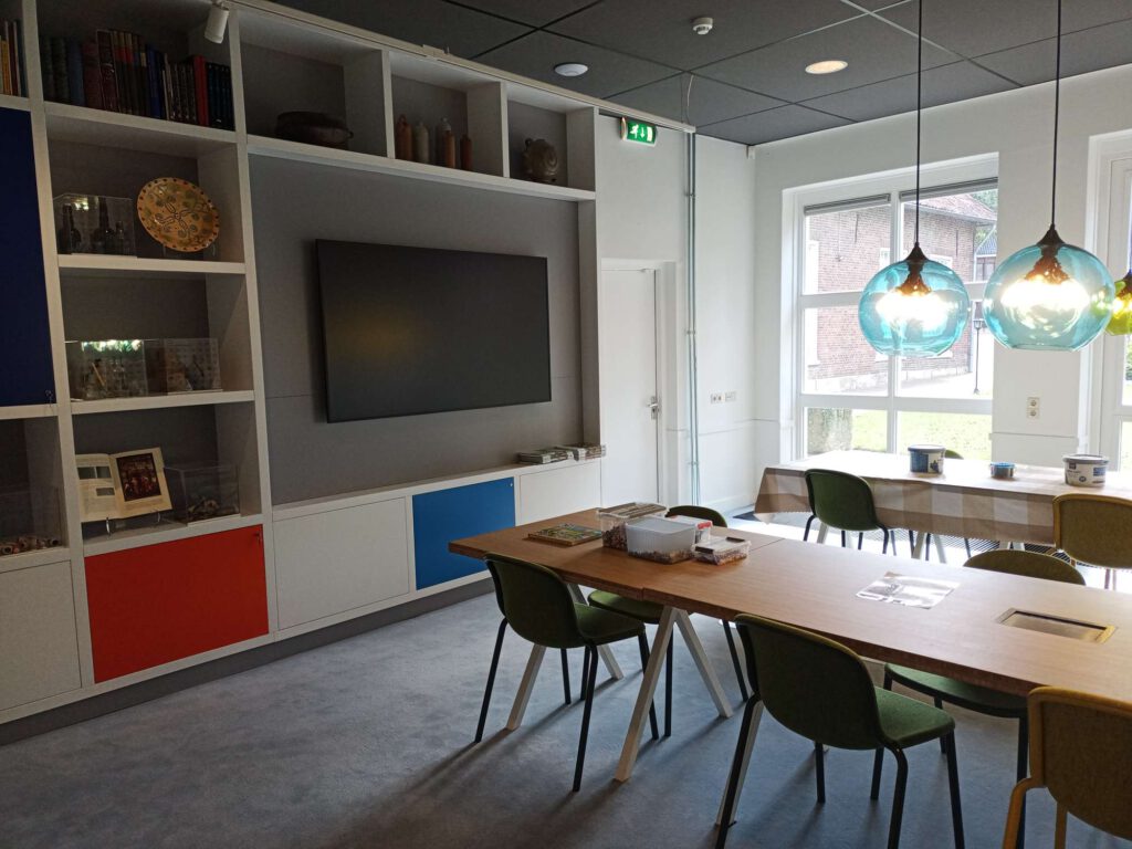 Een hoek van de studiezaal vanaf de muur gedraaid, je ziet nog wat tafels en kuipstoelen. De lampen zijn in een aquablauwkleur glas. Aan de wand hangt een televisie en er zijn verschillende objecten in de kast ernaast.