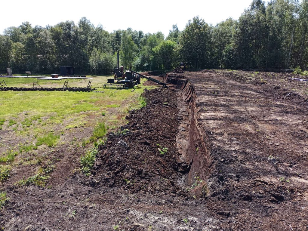 Rechts zie je bruine grond (veen), links, een stuk lager is nog gras te zien en halverwege het veld een turfpersmachine met een lopende band.