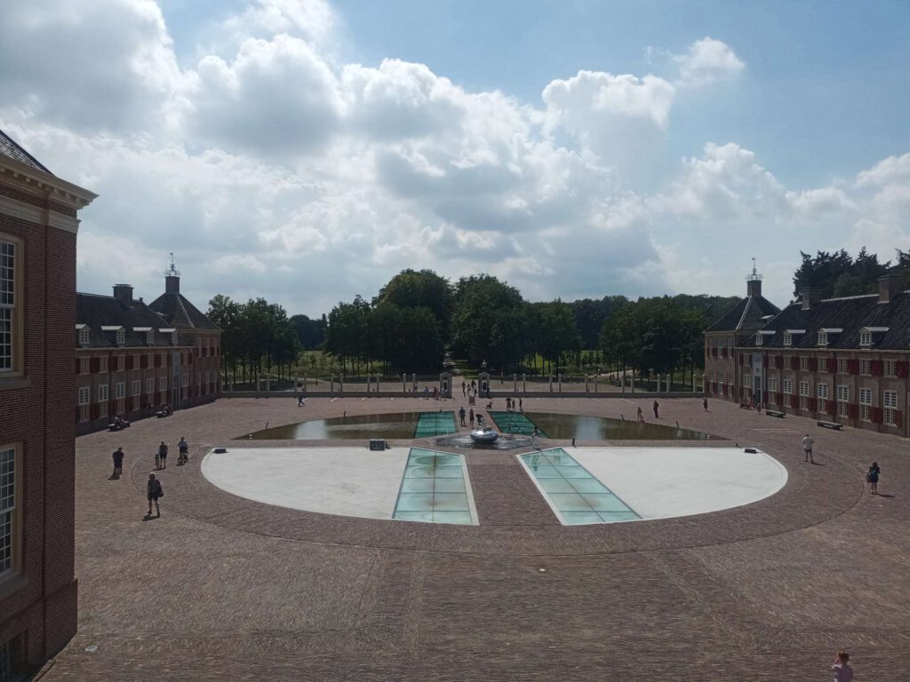 Links en rechts zijn vleugels te zien van het paleis. Op het voorplein zie je een soort ronde vorm met in et midden een soort fontein. Er lopen mensen over het plein.