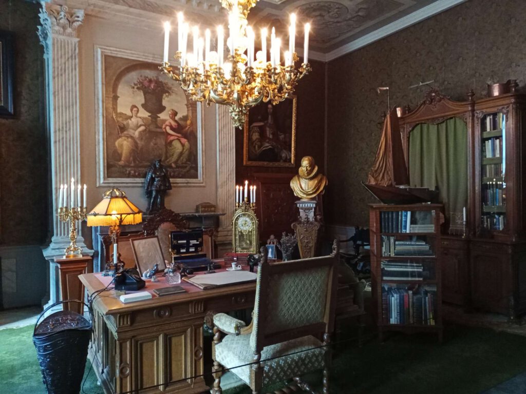 In de werkkamer hangt een kroonluchter, daaronder staat een donkerbruin bureau met houten stoelen, die bekleed zijn met een groene stof. Rondom het bureau staan verschillende zaken rondom schrijven, beelden en schilderijen.